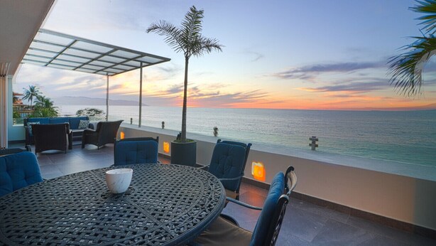 Eine Terrasse mit Meerblick bei Sonnenuntergang, mit einem runden Tisch, Stühlen und Kübelpflanzen.
