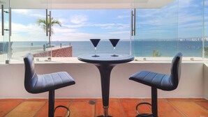 Ein Balkon mit Tisch und zwei Stühlen, der Blick auf die Küste mit klarem Himmel und dem Meer gewährt.