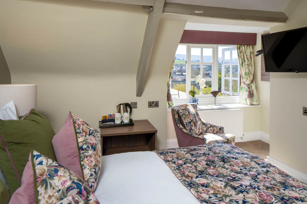 A bedroom with a bed, a chair, a table with a kettle, and a window with a view.