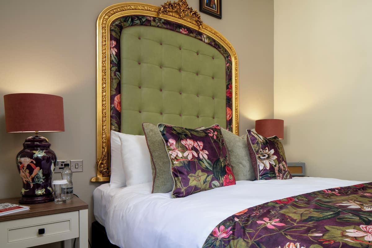 A hotel room with a large bed, two bedside tables, a nightstand, a decorative vase, and a wall-mounted picture.