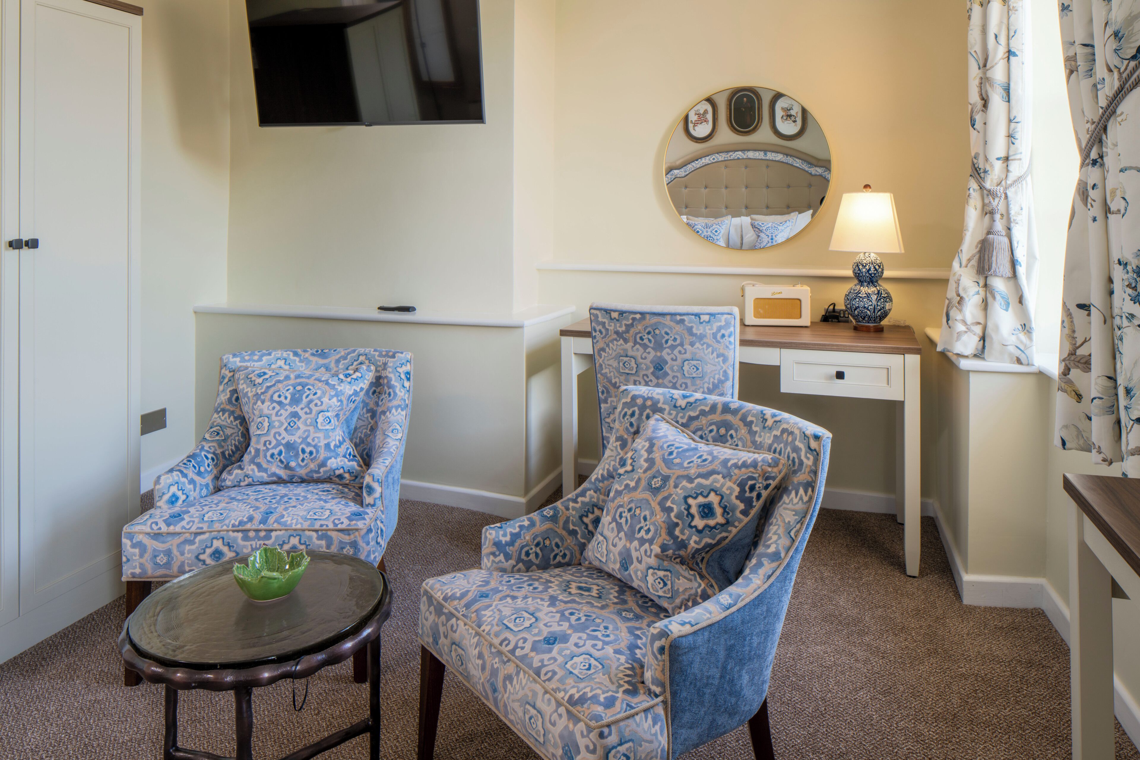 A room with two patterned armchairs, a small table with a green bowl, a bedside table with a lamp, a mirror, and a TV mounted on the wall.