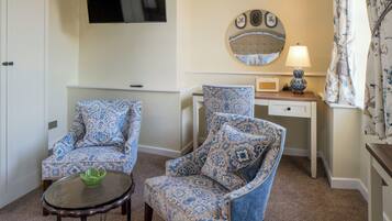 A room with two patterned armchairs, a small table with a green bowl, a bedside table with a lamp, a mirror, and a TV mounted on the wall.