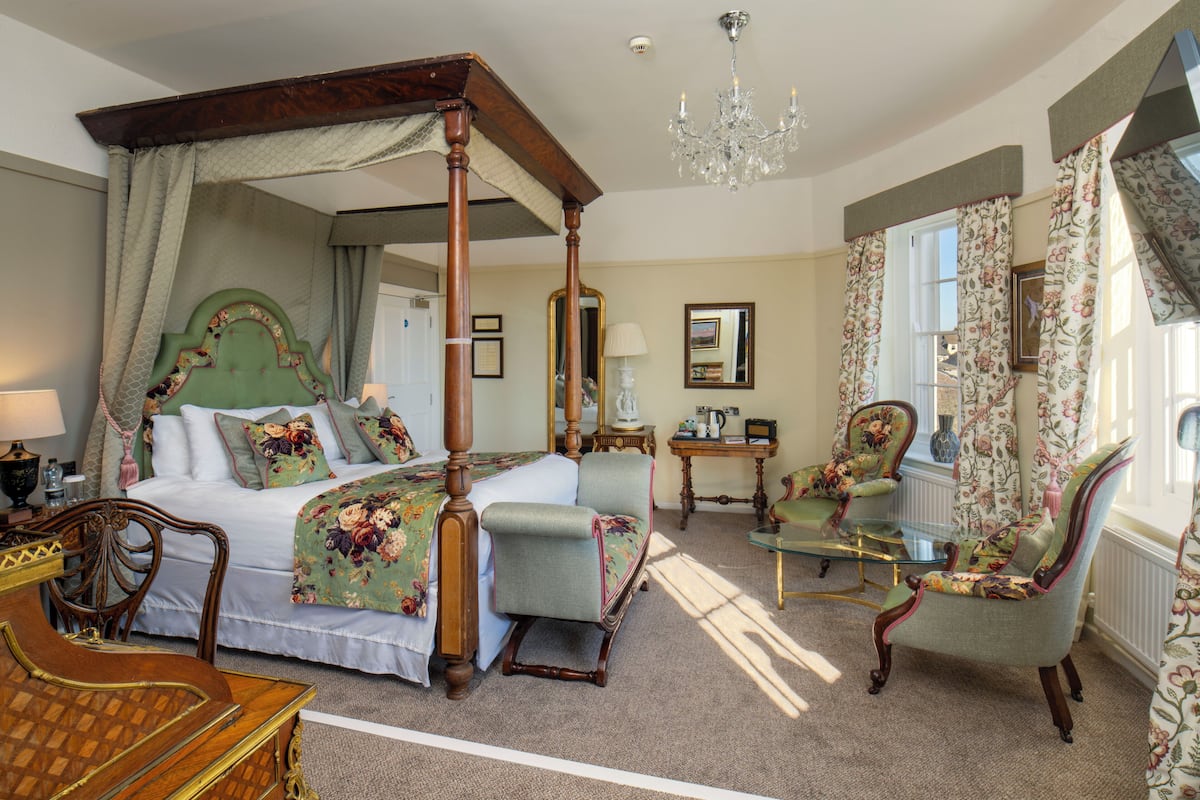 A four-poster bed with a canopy, a sitting area with a glass table, and a chair with floral upholstery.