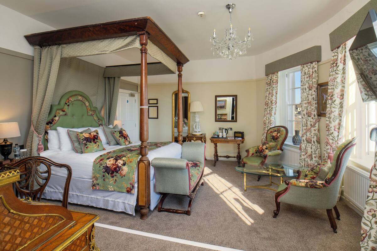 A four-poster bed with a canopy, a sitting area with a glass table, and a chair with floral upholstery.