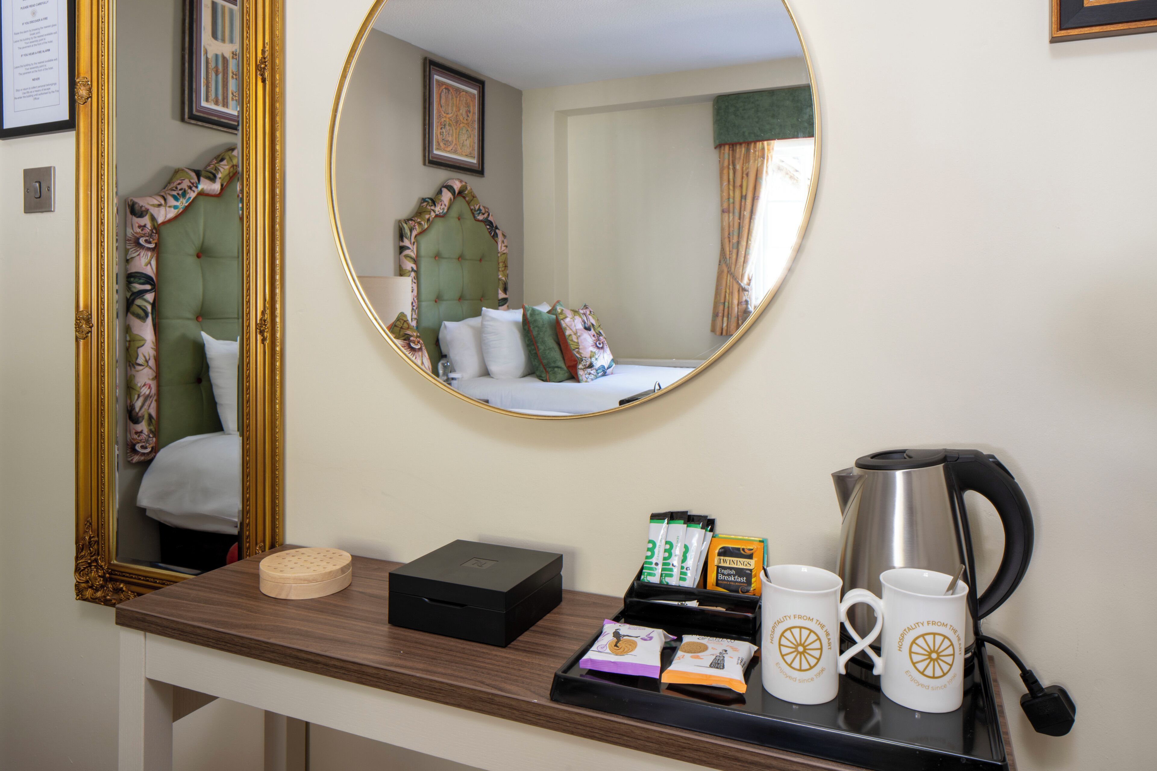 Quarto de hotel com cama, espelho, mesa de cabeceira com chaleira, bandeja com sachês de café e uma caixa.