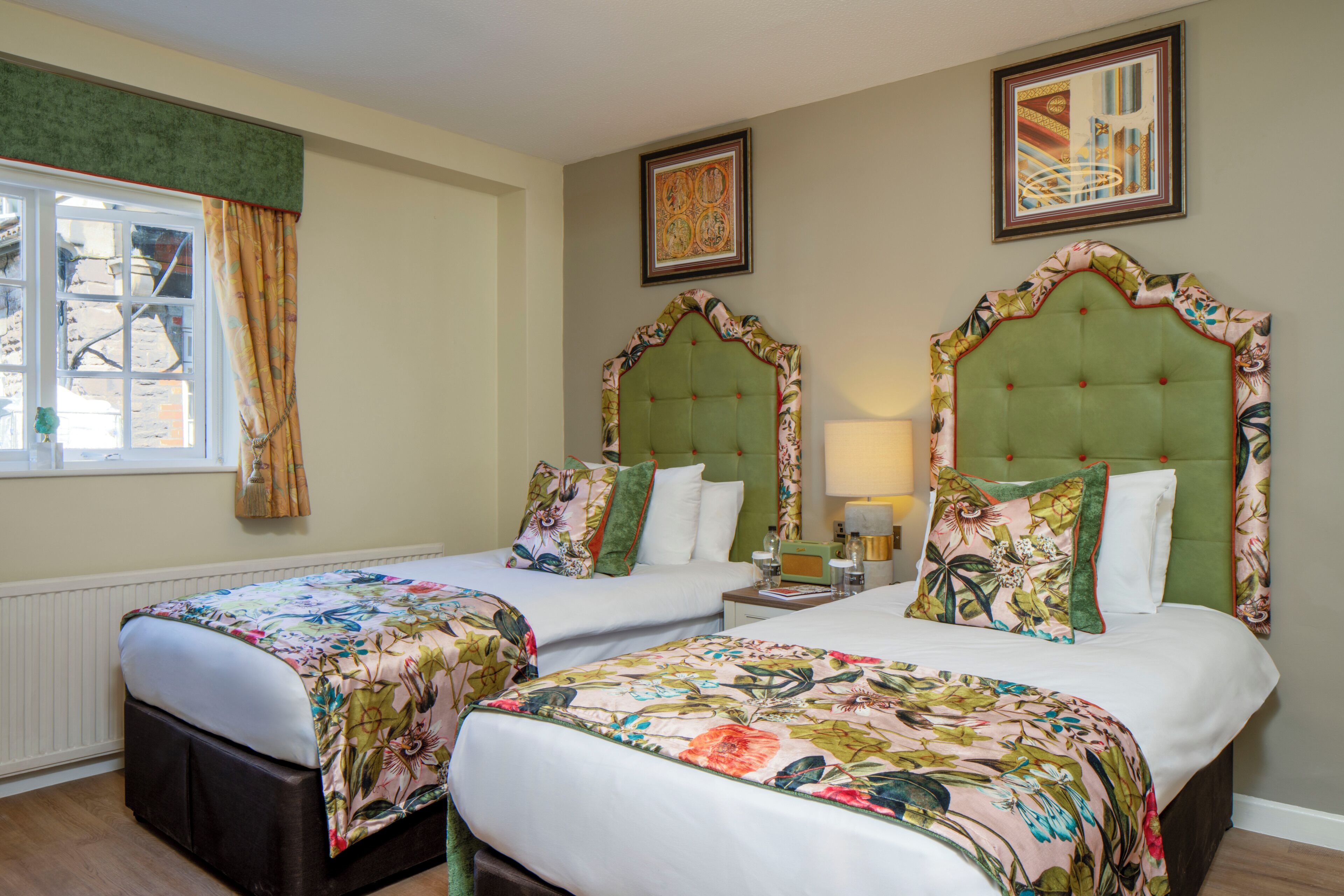 A hotel room with two beds, a window with curtains, a framed picture on the wall, and a bedside table with a lamp.