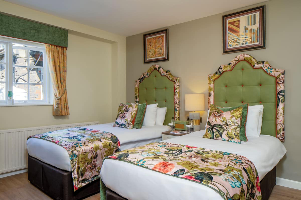 A hotel room with two beds, a window with curtains, a framed picture on the wall, and a bedside table with a lamp.