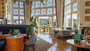 Ein Hotelzimmer mit großem Fenster, einer Sitzecke, einem Esstisch mit Stühlen und Blick auf die Landschaft.