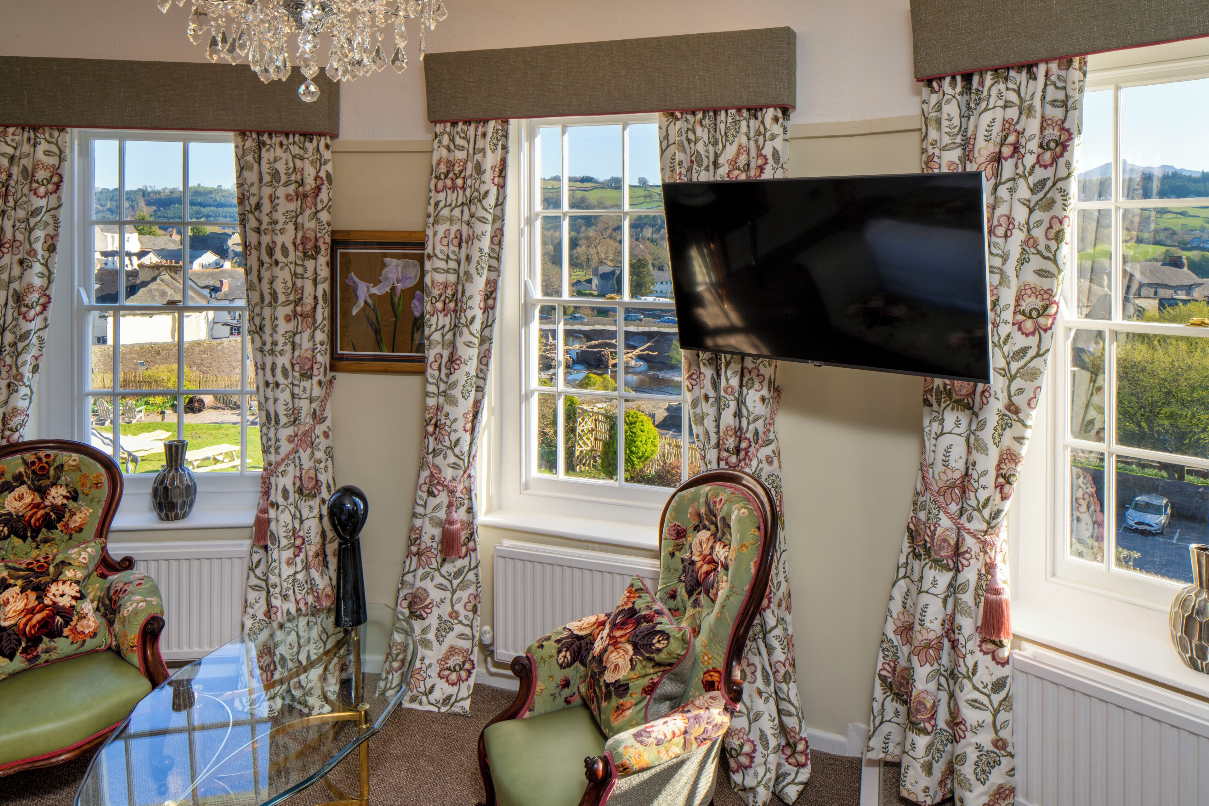 Una habitación con una televisión montada en la pared, dos sillones con estampado floral, una mesa de cristal y vistas al campo a través de amplios ventanales.