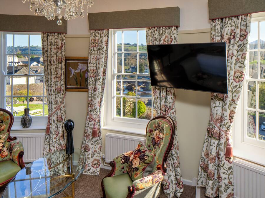 Una habitación con una televisión montada en la pared, dos sillones con estampado floral, una mesa de cristal y vistas al campo a través de amplios ventanales.
