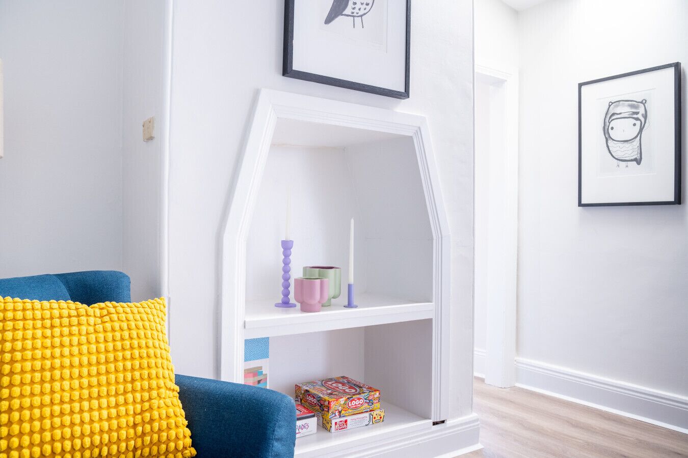 A room with a white arched bookshelf, a blue chair with yellow cushions, and framed artwork on the walls.