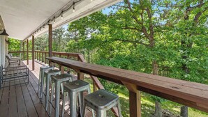 A covered outdoor deck with metal stools and a wooden table, surrounded by trees. - Lakeside Hideaway, Fire-Pit | Near Marina (Rogers)