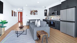 A modern living room with a grey sofa, a coffee table, and a small wooden side table. A kitchen with dark cabinets and stainless steel appliances is visible. - Modern Hill Country Condo for 4 w/ Resort Perks! (New Braunfels)