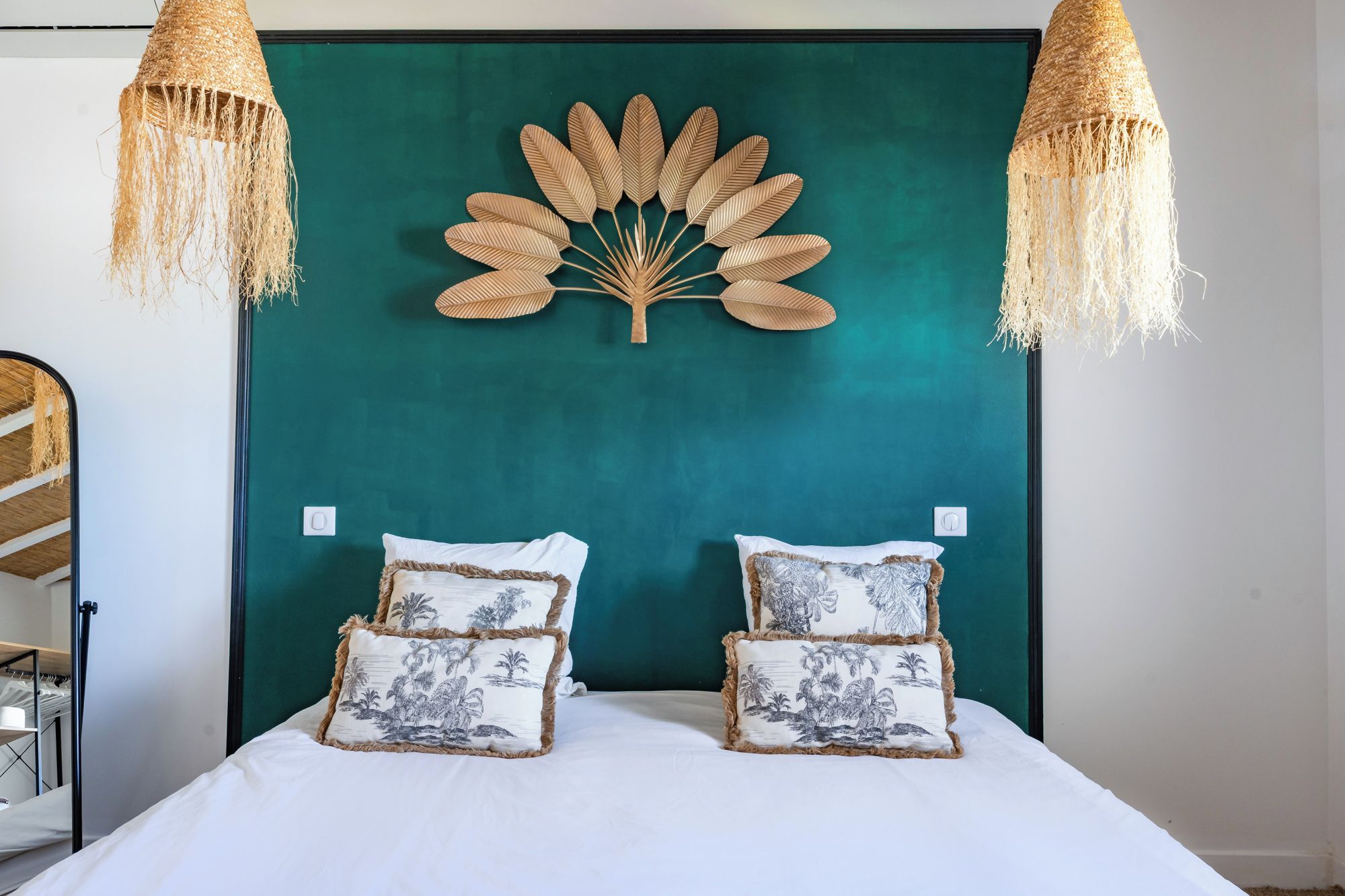Un lit avec une literie blanche et deux coussins décoratifs à motifs tropicaux, une tête de lit de couleur turquoise et deux lampes suspendues ornées de glands.