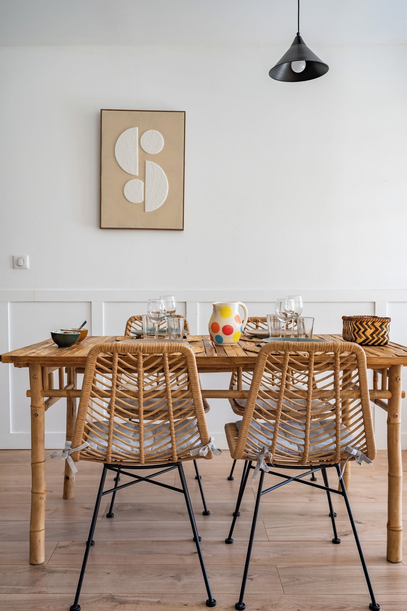 A dining area with a wooden table, wicker chairs, a colorful mug, and a pendant light.