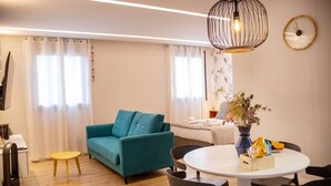 Ein modernes Wohnzimmer mit einem türkisfarbenen Sofa, einem runden Esstisch mit Stühlen, einer Pendelleuchte und einer Wanduhr.