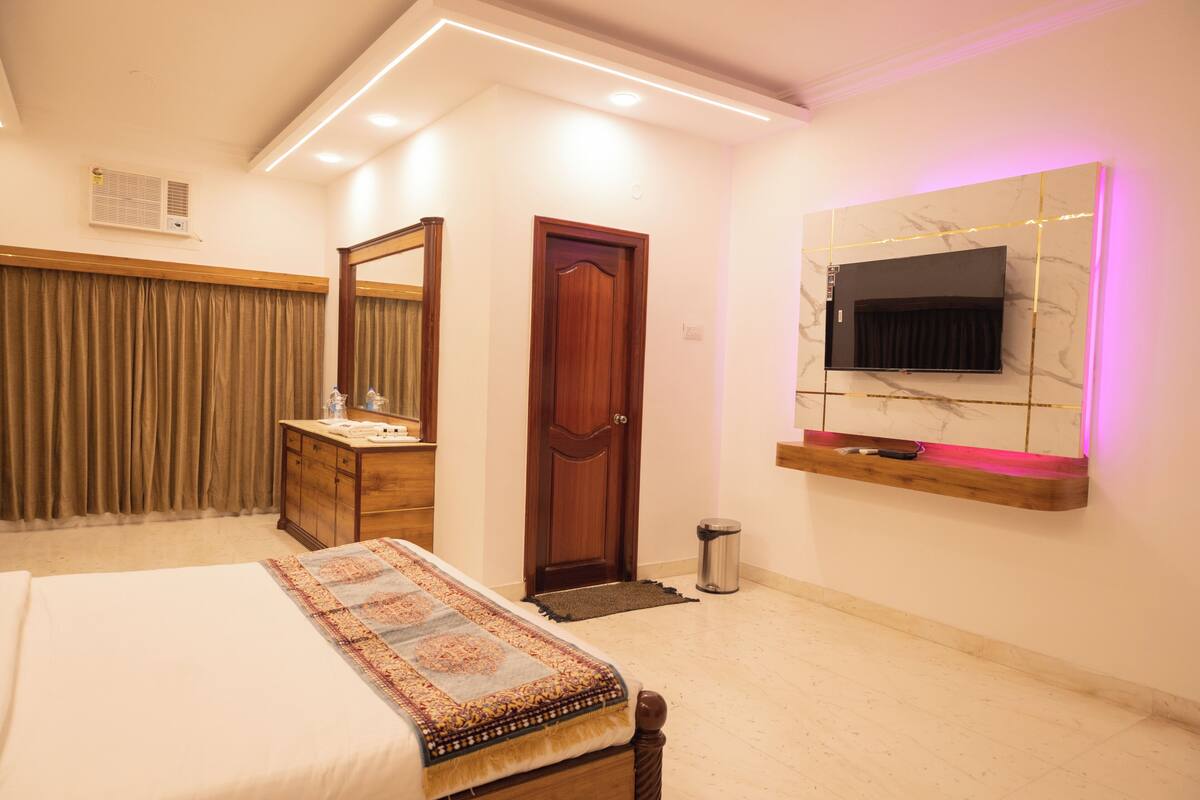 A hotel room with a bed, a wooden dresser, a mirror, a TV mounted on the wall, and a door.
