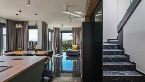 Un hall d’hôtel moderne avec un comptoir d’accueil en bois, un espace salon avec un canapé et des fauteuils, un escalier avec des marches en marbre noir et de grandes fenêtres avec des rideaux.