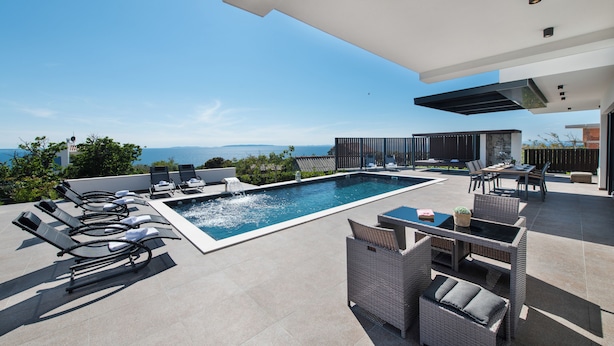 Un espace piscine extérieur moderne, doté de chaises longues, d’un ensemble de mobilier de jardin et offrant une vue sur la mer.