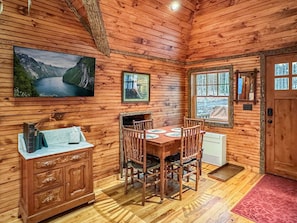 A wooden cabin interior with a dining table, chairs, a cabinet, a fireplace, and a window with a view. - Lakeside Lodge on 7th Lake - The Carriage House (Inlet)