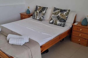 Una cama bien hecha con un edredón blanco, dos almohadas con estampado de hojas y una toalla doblada al lado. Hay dos mesitas de noche de madera con jarrones decorativos sobre ellas.