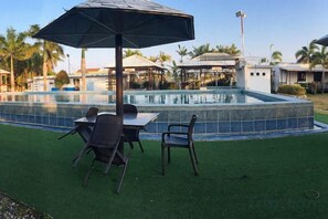 Zona de piscina exterior con mesa y sillas, rodeada de césped artificial.
