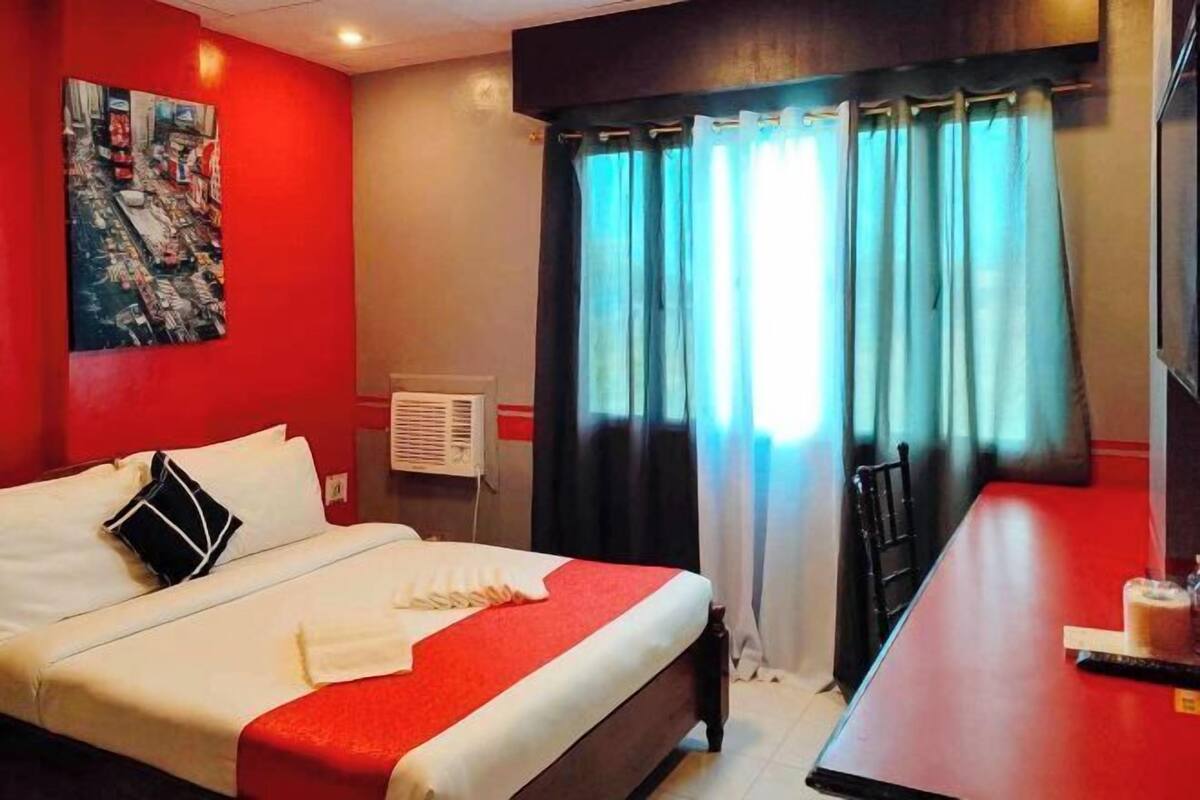 A hotel room with a bed, a desk, a painting on the wall, and a window with curtains.