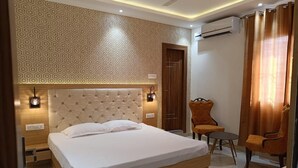 A bedroom with a large bed, a wooden headboard, two bedside tables, a chair, a small table, and an air conditioning unit. - Cozy hotel in terrific Purnia with WiFi, AC (Purnia)