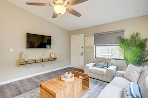 A living room with a ceiling fan, a mounted TV, a wooden coffee table, a sofa, a chair, a window with a view, and a plant.