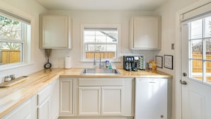 A kitchen with white cabinets, a wooden countertop, a built-in dishwasher, and a window with a view of a wooden fence.