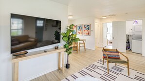 A modern living room with a flat-screen TV, a wooden console table, a potted plant, a dining area with a wooden chair, and a kitchen in the background.