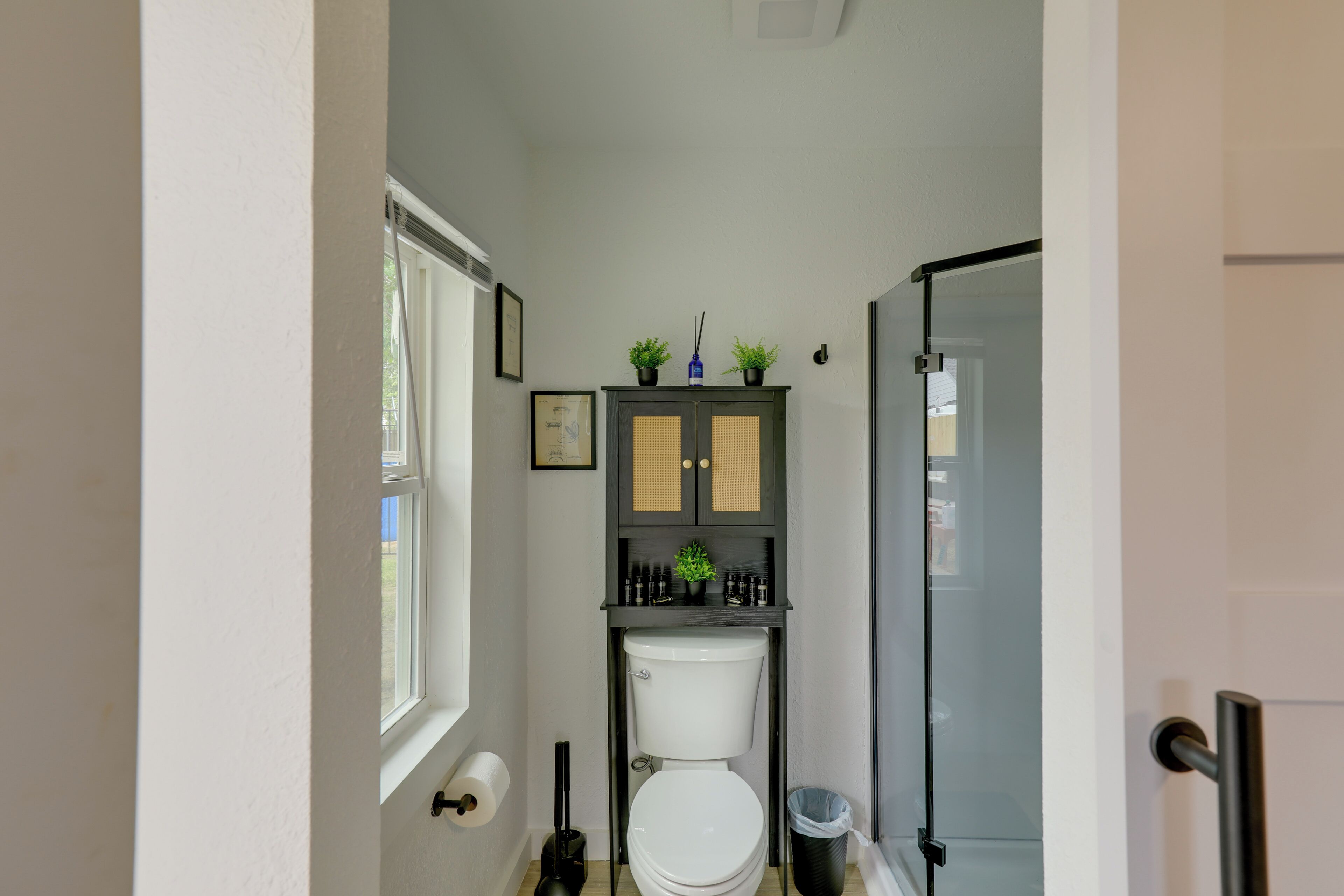 Une petite salle de bain comprenant des toilettes, un meuble avec des plantes, une poubelle et une porte.