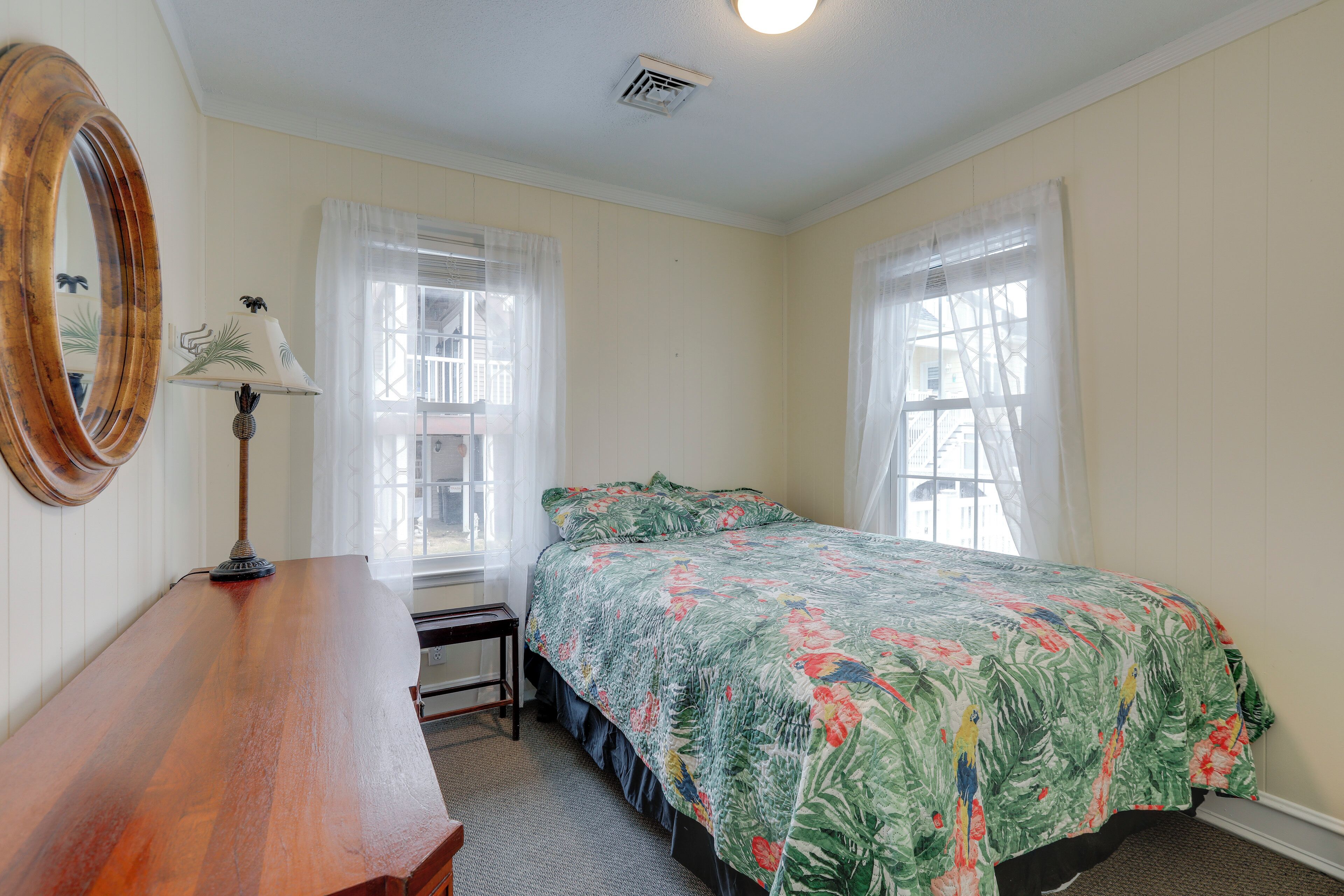 A bedroom with a bed, a wooden dresser, a lamp, and two windows with sheer curtains.