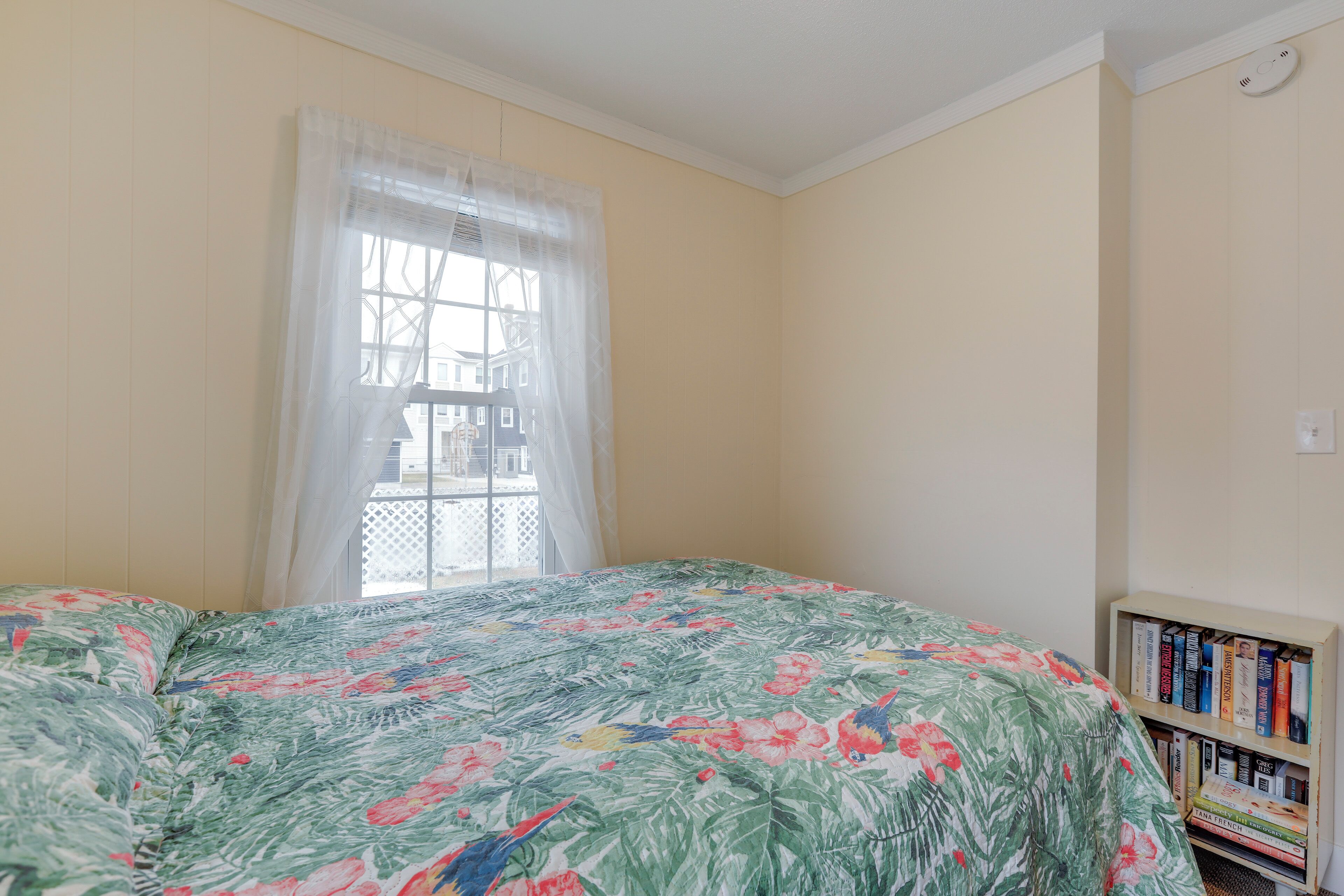 A room with a bed, a window with sheer curtains, and a bookshelf with books.
