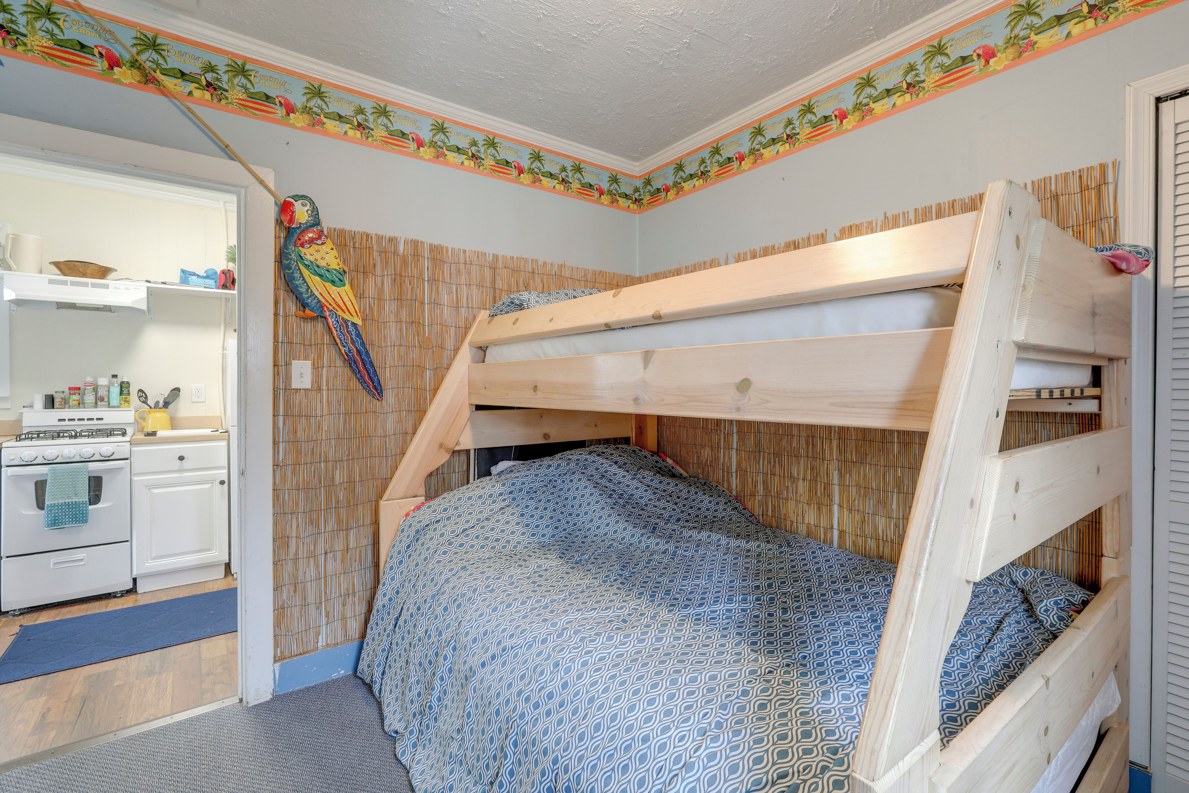 A bunk bed with a patterned bedspread, a kitchen area with white appliances, and a decorative parrot on the wall.