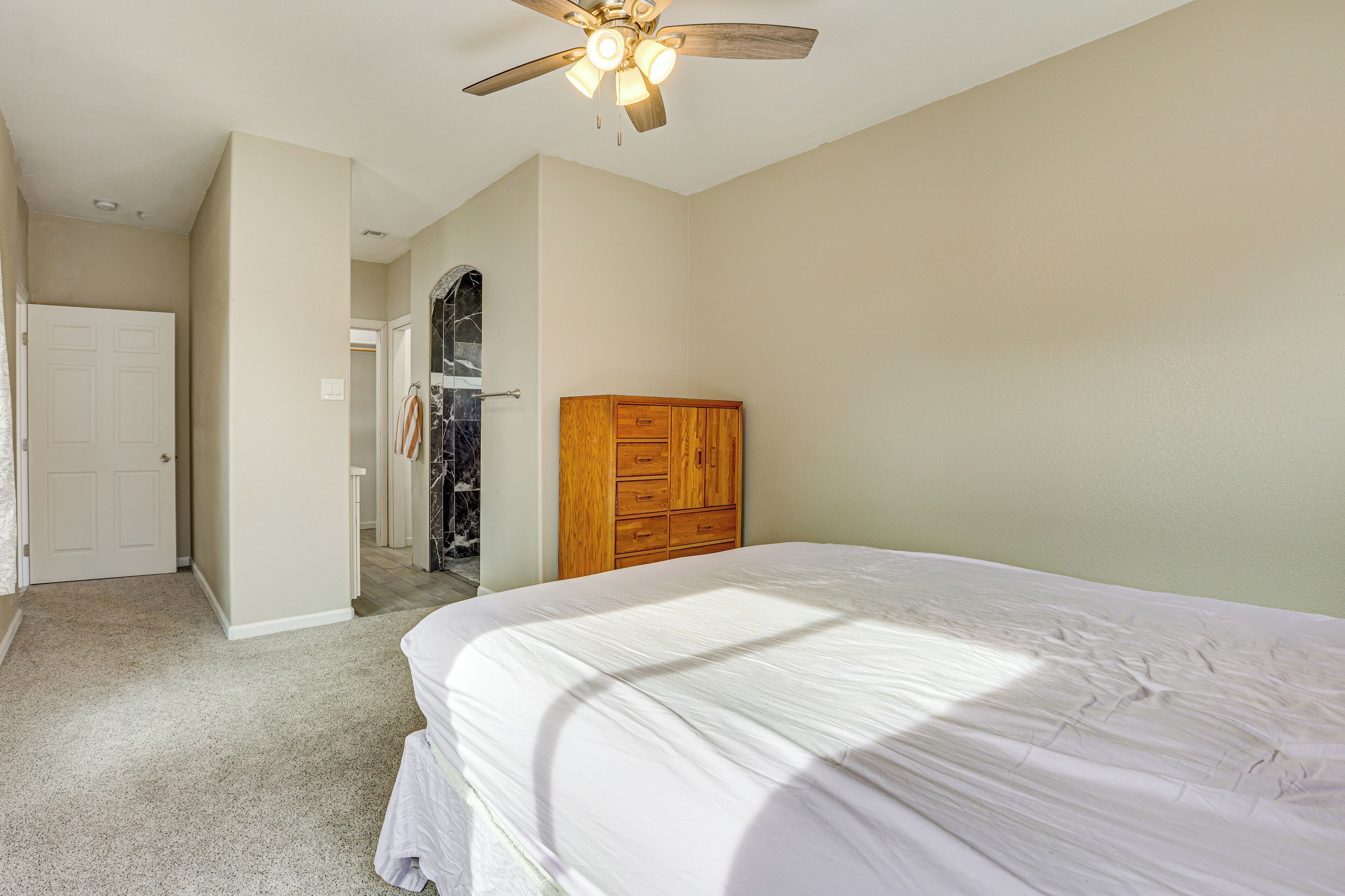 Um quarto com uma cama, cômoda de madeira, ventilador de teto e porta que dá para um corredor.