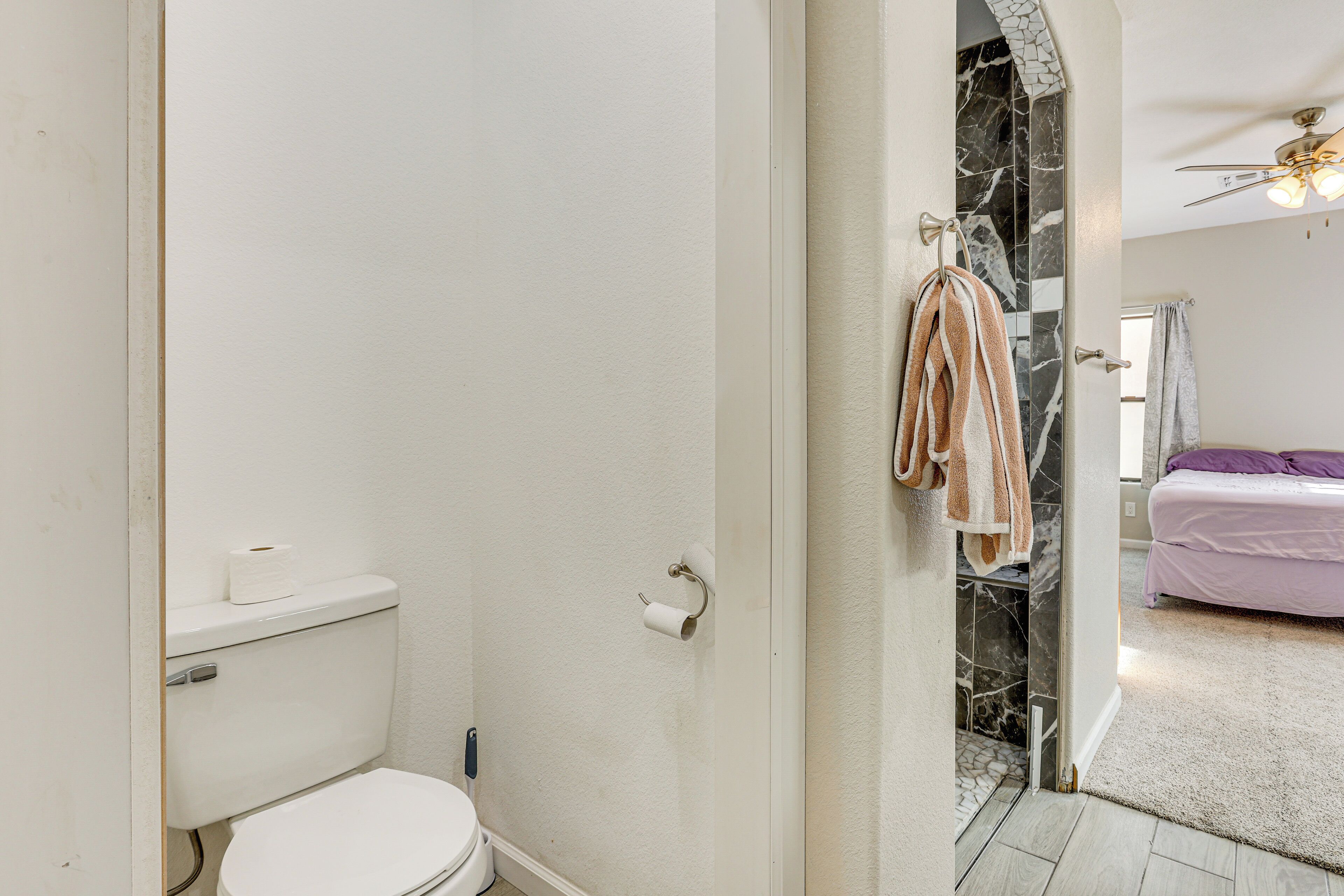 Un baño con inodoro, un perchero con toallas, una zona de ducha con pared de mármol, y una cama con colcha morada en la habitación contigua.