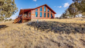 A wooden cabin with a large deck and multiple windows, surrounded by dry grass and trees.