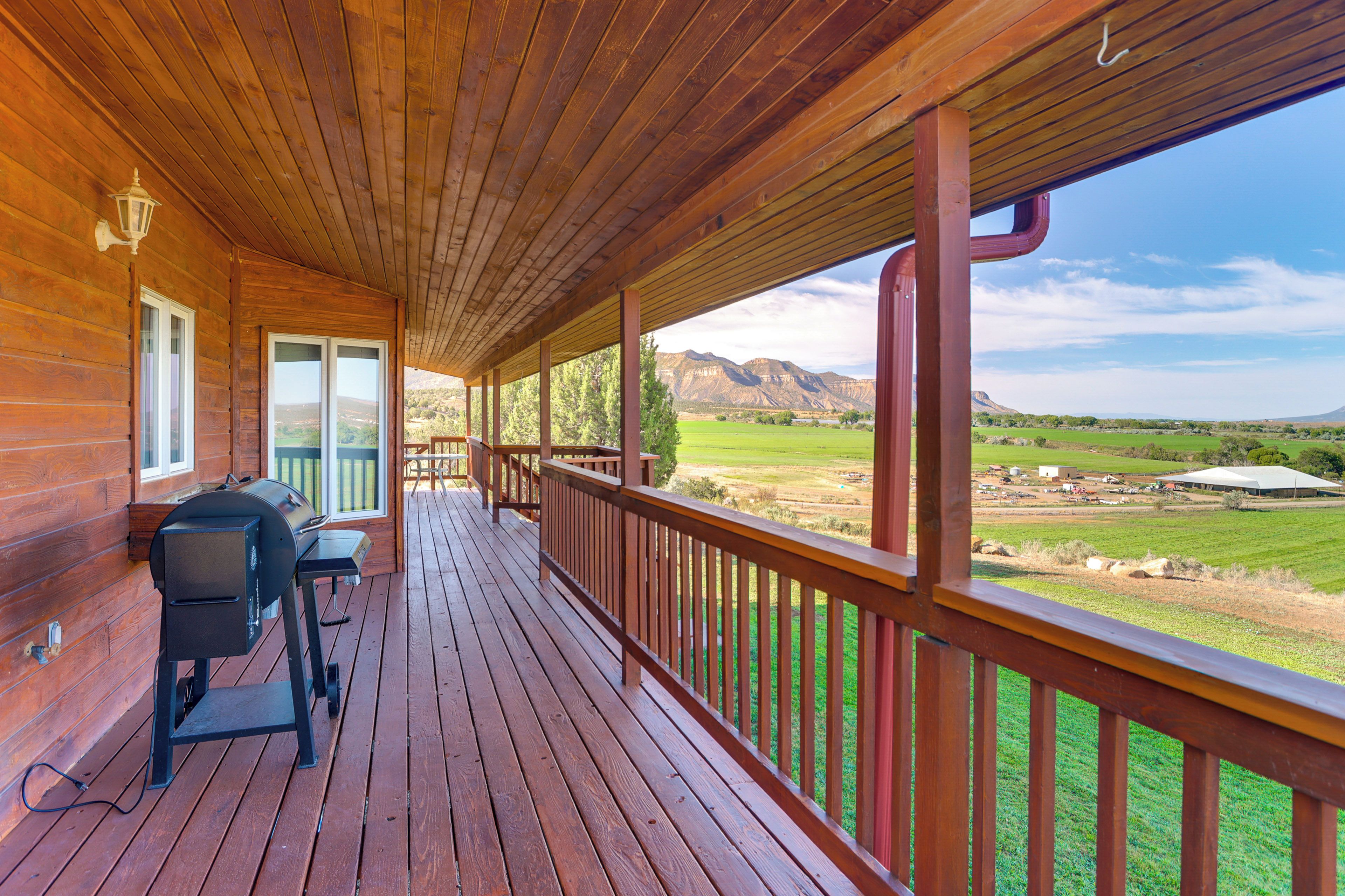 A wooden deck with a grill and seating area, overlooking a landscape with mountains and fields.