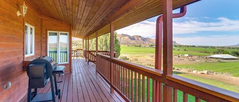 A wooden deck with a grill and seating area, overlooking a landscape with mountains and fields.