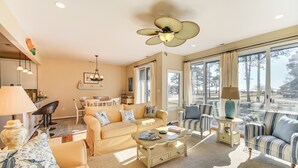 Ein geräumiges Wohnzimmer mit einem beigen Sofa, einem gestreiften Sessel und einem Couchtisch. Es gibt einen Deckenventilator, einen Essbereich und große Fenster mit Blick ins Freie.