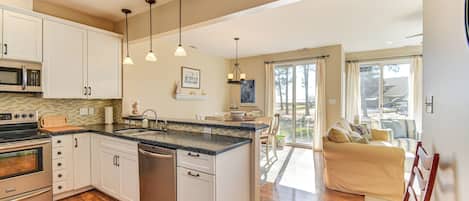 A modern kitchen with white cabinets, a stainless steel oven, and a dishwasher. There is a dining area with a yellow sofa and a dining table.