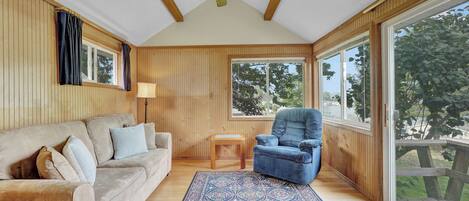 A cozy room with a wooden ceiling, a large window, a sofa, a chair, and a patterned rug.