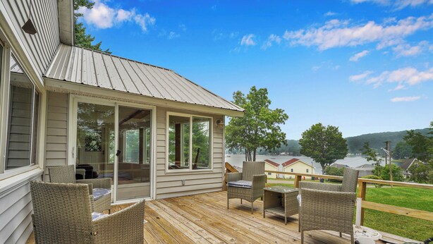 A wooden deck with wicker furniture, a covered area with glass doors, and a view of a lake and houses in the background. - Private Dock & Lake Views: Home on Lime Lake (Machias)