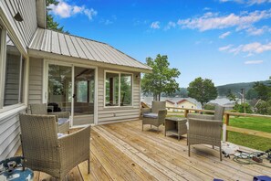 A wooden deck with wicker furniture, a covered area with glass doors, and a view of a lake and houses in the background. - Private Dock & Lake Views: Home on Lime Lake (Machias)