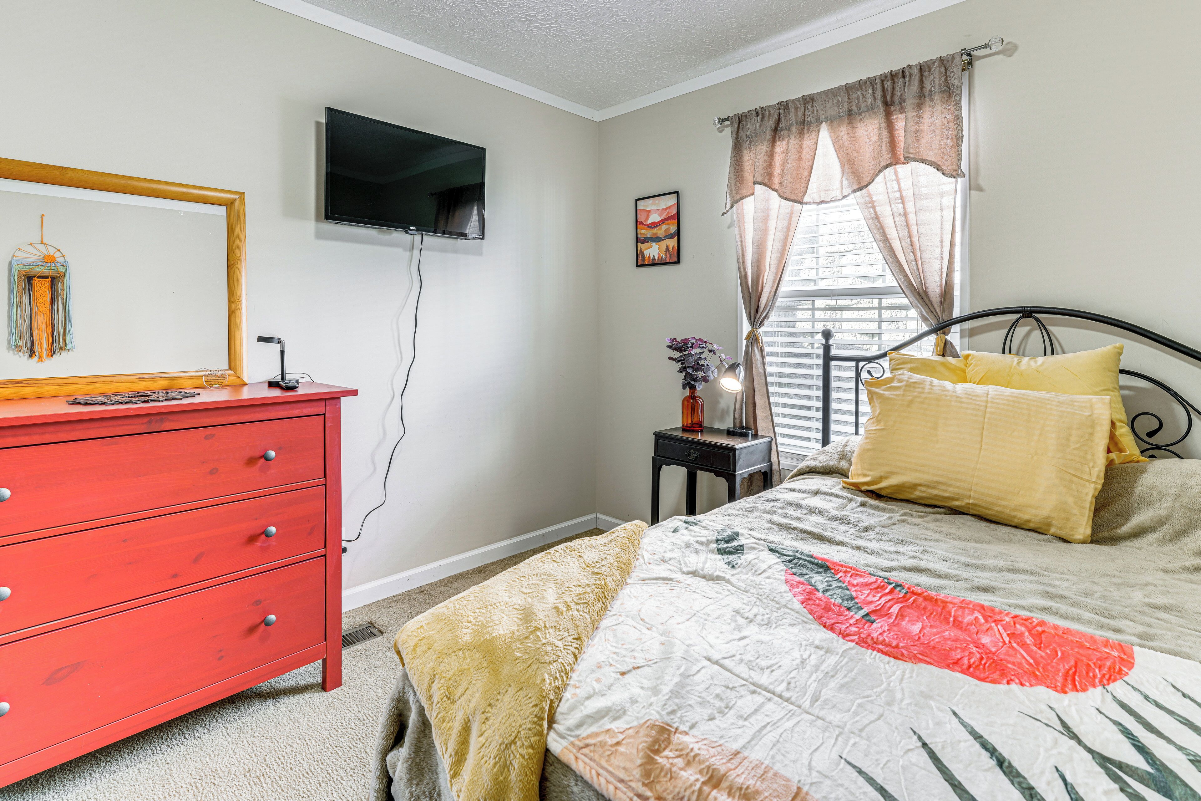 A bedroom with a red dresser, a bed with a patterned bedspread, a television mounted on the wall, and a window with curtains.