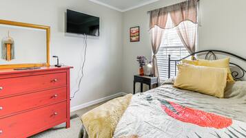 A bedroom with a red dresser, a bed with a patterned bedspread, a television mounted on the wall, and a window with curtains.