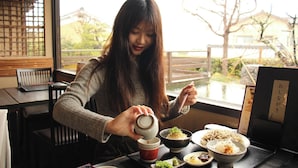 Una persona sirve té en una taza sobre una mesa con diversos platos, entre ellos fideos y un tazón de sopa, en una habitación con ventana y un separador tradicional.