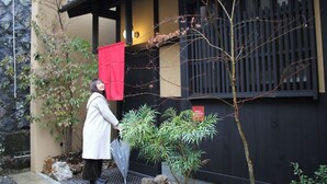 Una persona de pie junto a un edificio con fachada negra y beige, un paño rojo colgando y un pequeño árbol en primer plano.
