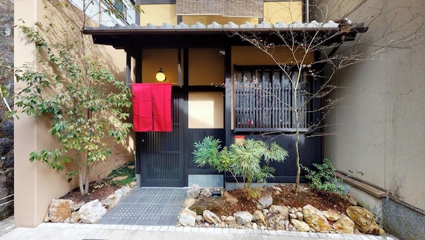 Una casa tradicional japonesa con entrada negra, una toalla roja colgada en la puerta y un pequeño jardín con rocas y plantas.